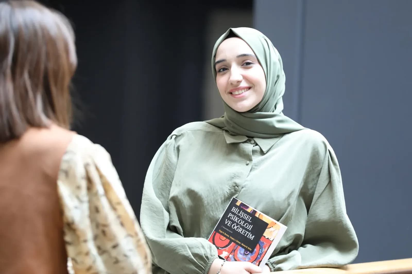 A young woman in a headscarf smiles while holding a book, indoors.