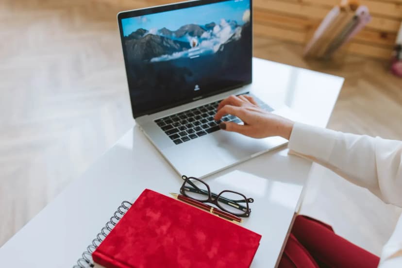 A person working on a laptop with a red notebook and glasses on a white table.