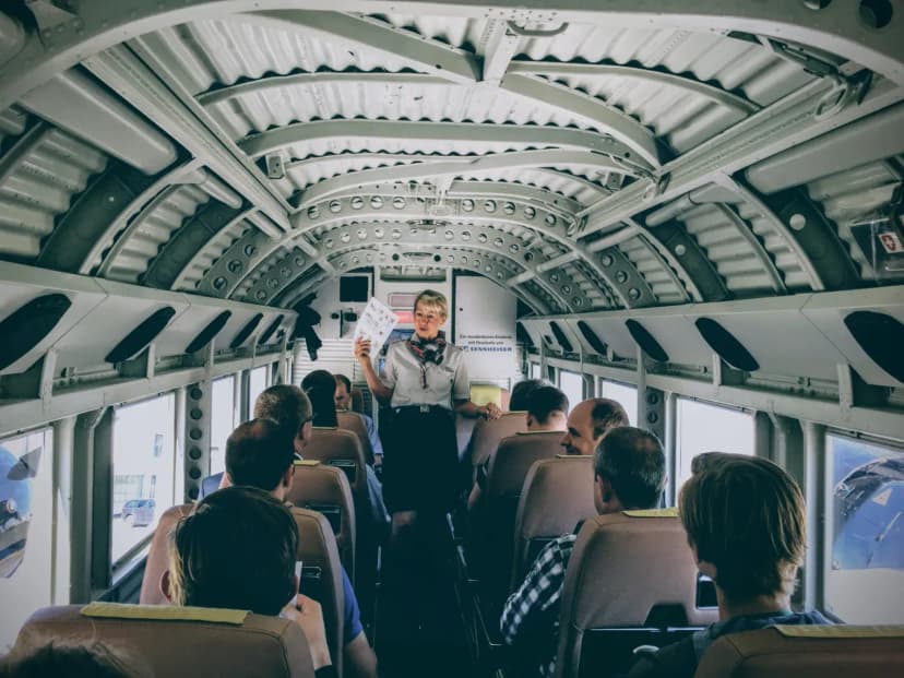 Inside a vintage aircraft, a flight attendant gives safety instructions to passengers.