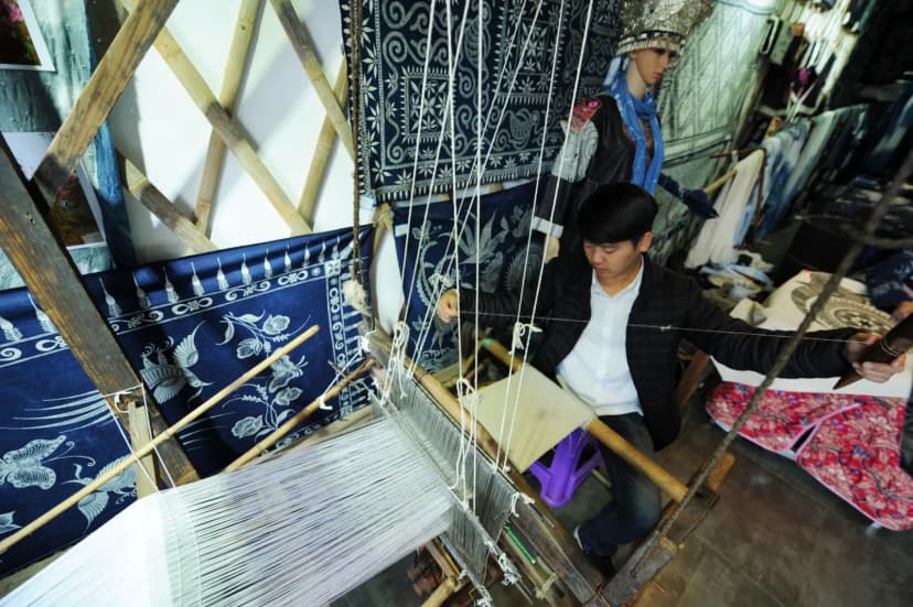 Young man weaving on a loom in a traditional workshop with intricate fabric designs.