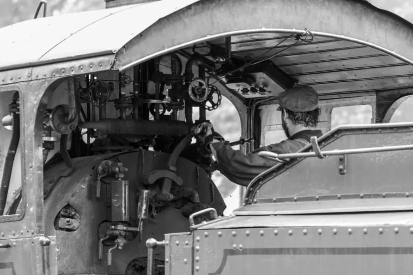 A vintage train engineer works in the steam locomotive's cabin, captured in black and white.