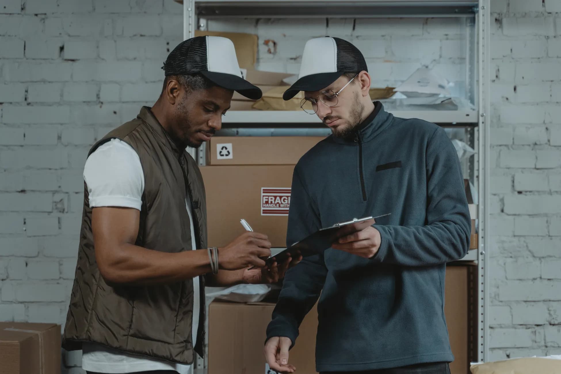 Two male couriers discussing and signing a clipboard in a warehouse setting, surrounded by packages.