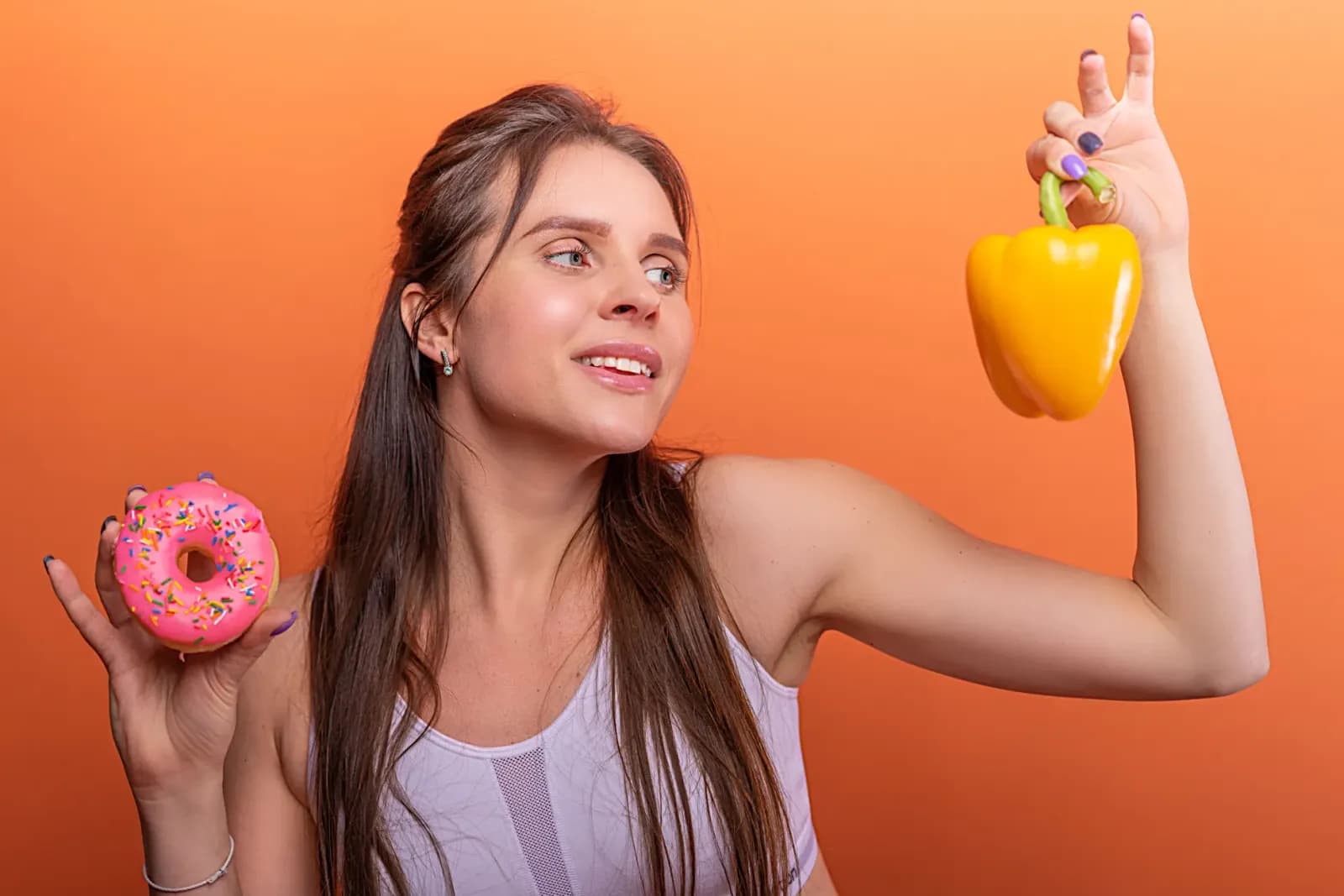 Woman in sports bra juggling choice between a donut and bell pepper on orange background.