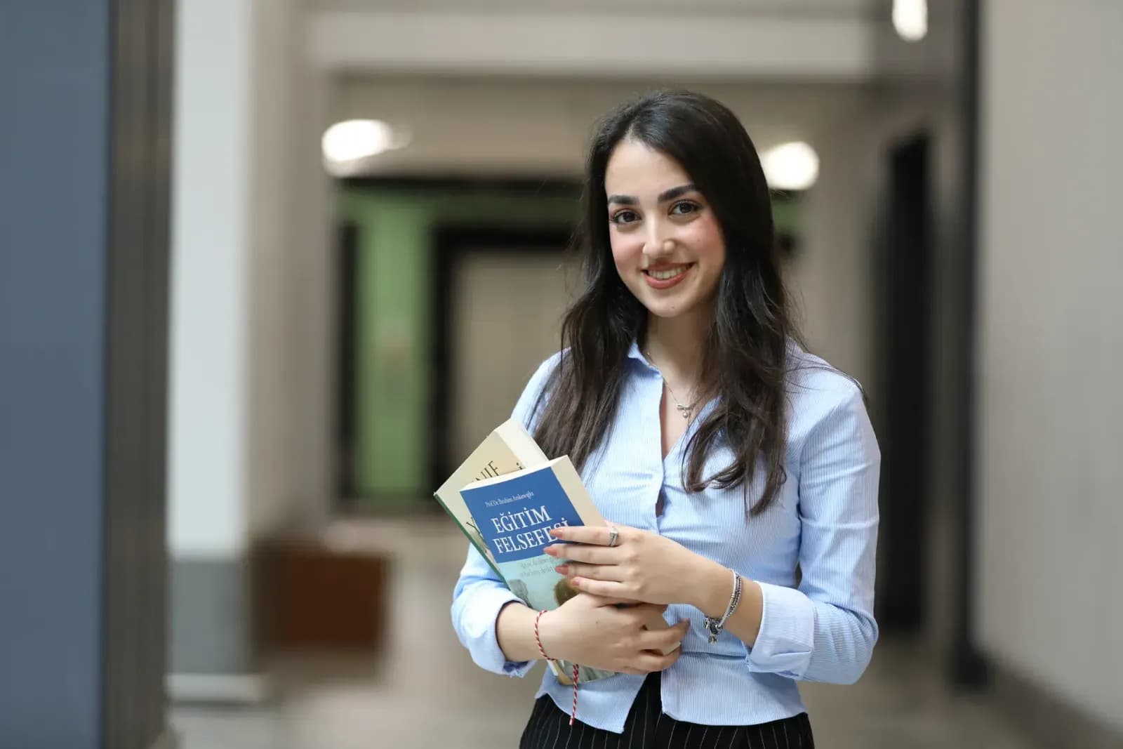 Young woman with books, standing in a hallway, smiling confidently.