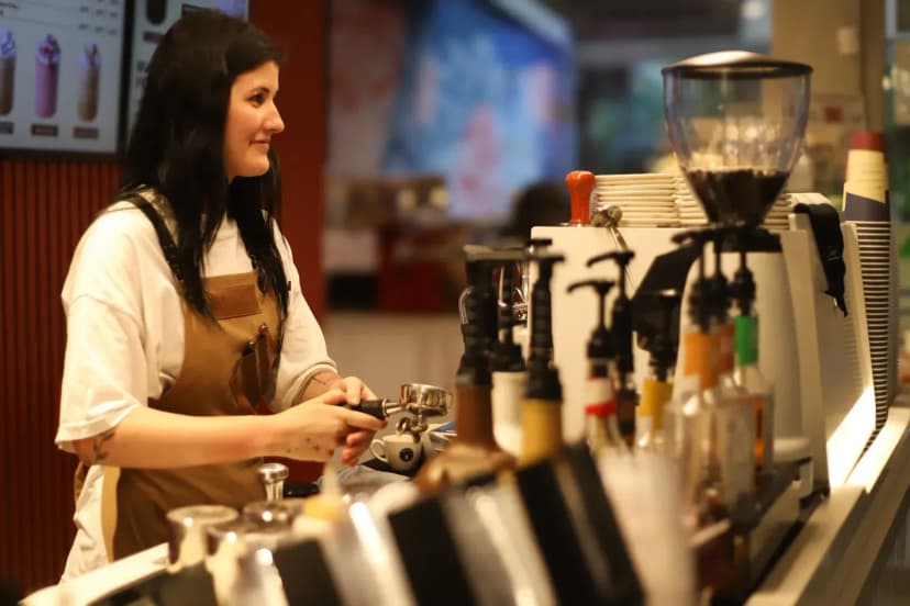 Barista in an apron skillfully making coffee in a bustling café environment with a variety of syrups and equipment.