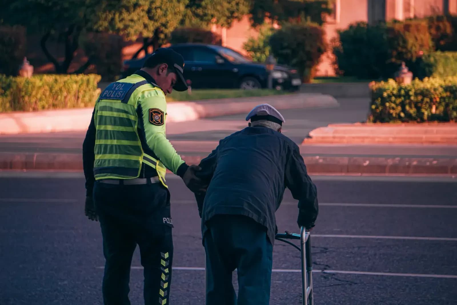 A police officer helps an elderly man with a walker cross the street at sunset.
